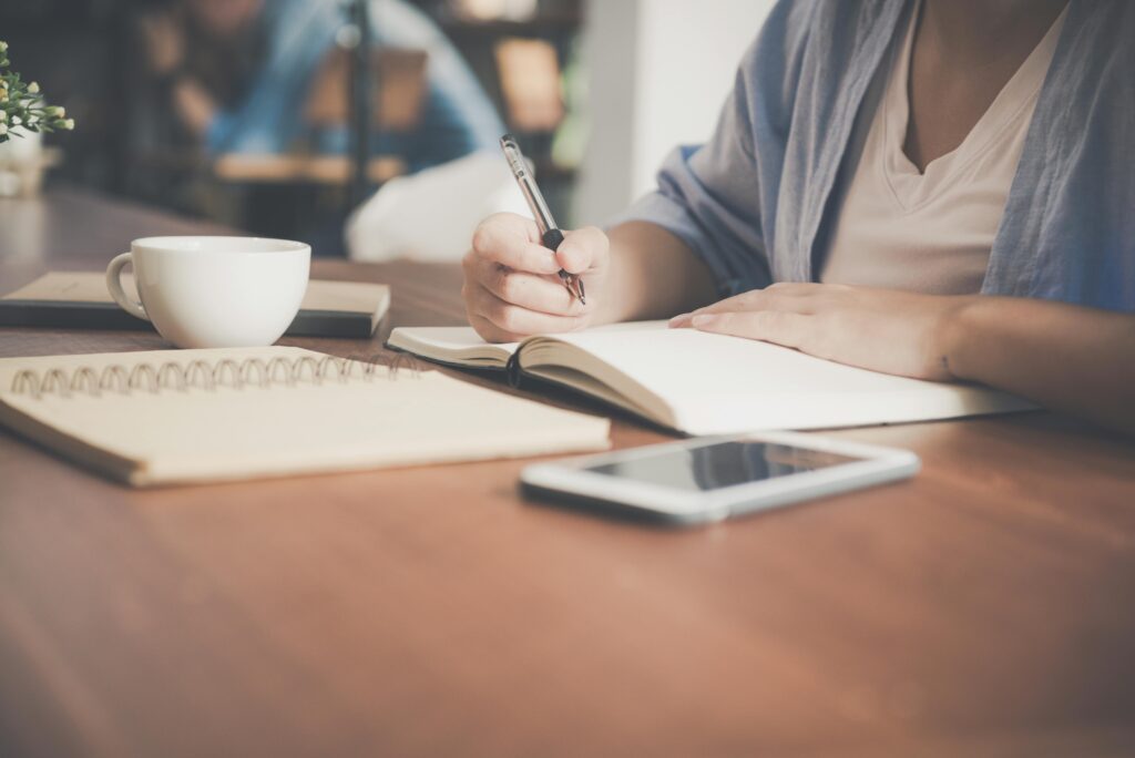 pexels-photo-733856-733856 A woman writes in a notebook at a café table with a coffee and smartphone nearby.